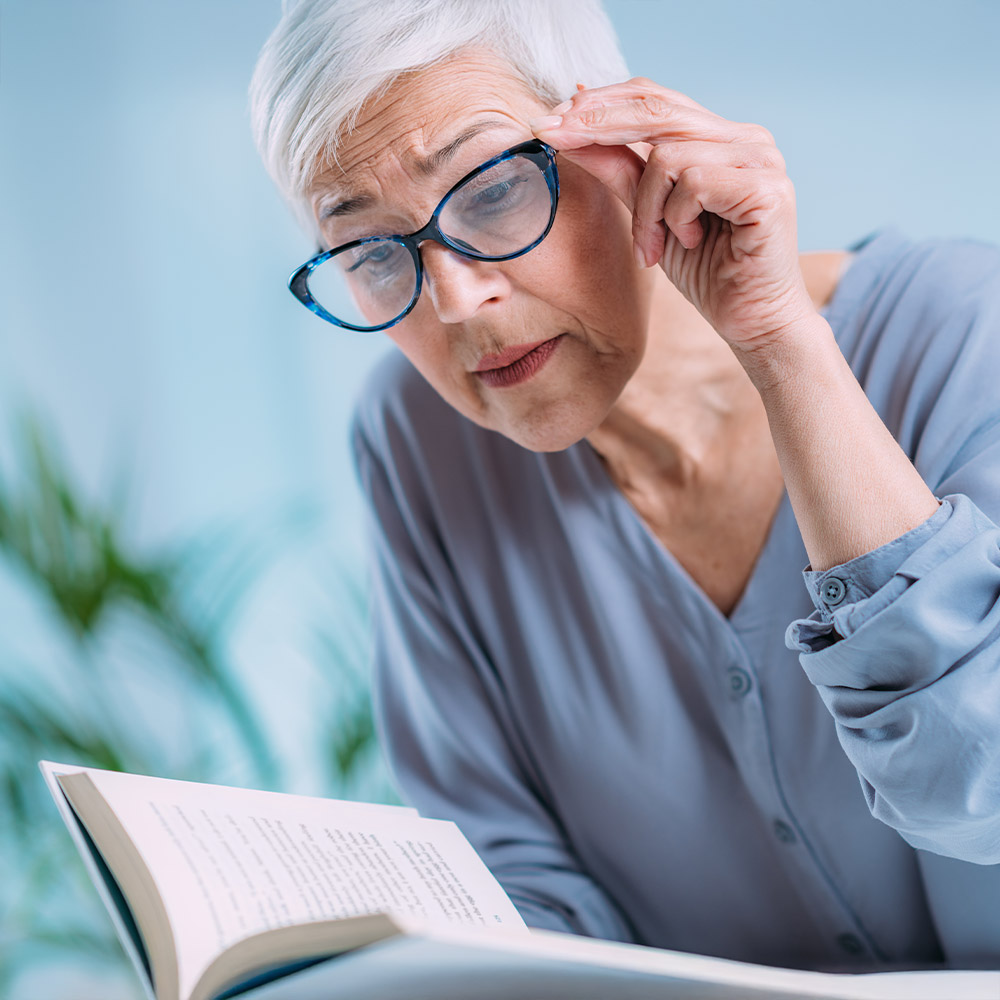 A senior woman squinting and adjusting her glasses while reading a book, illustrating common cataract symptoms like blurry vision and difficulty reading.