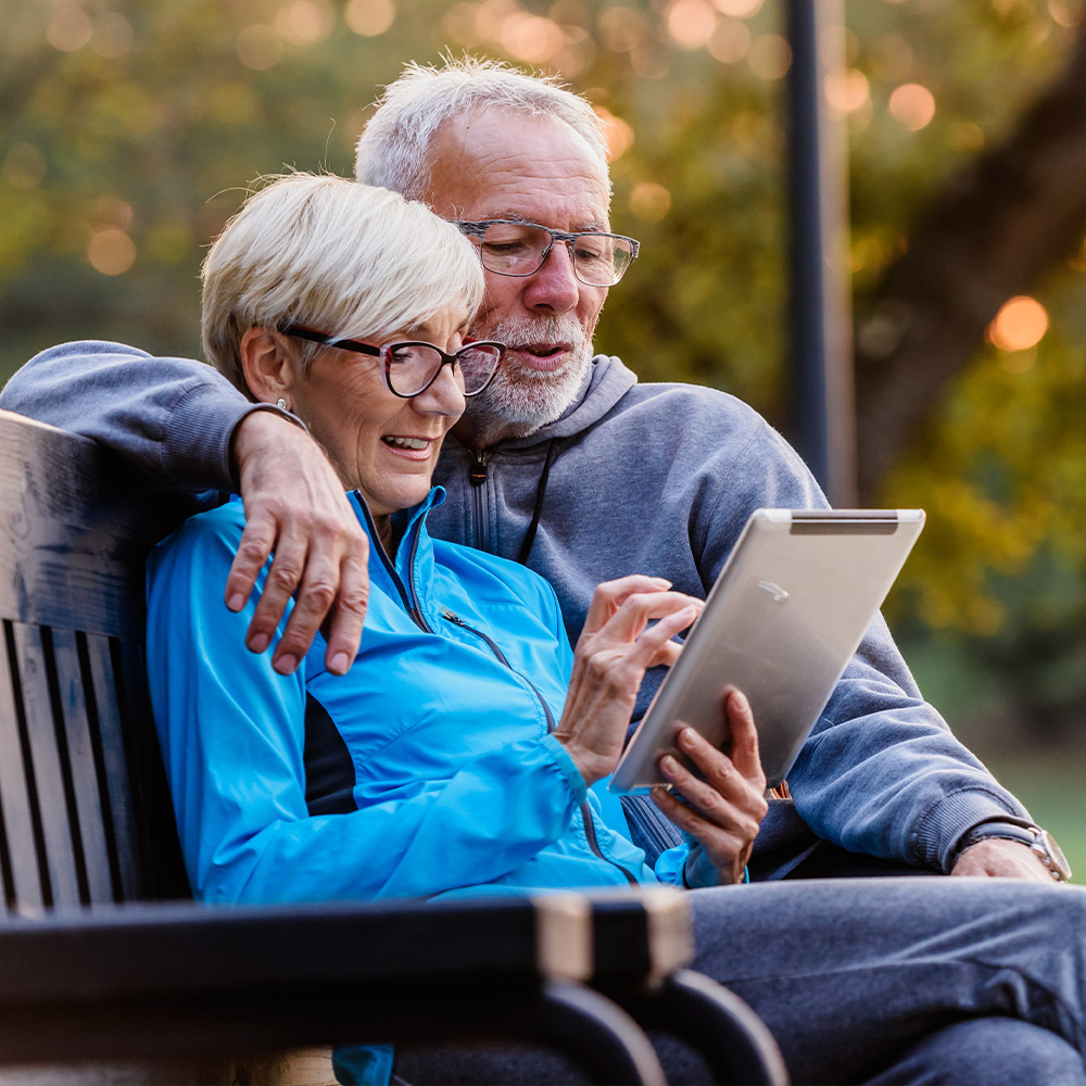 Senior couple in glasses using a tablet outdoors, showing clear vision for digital devices after cataract surgery.
